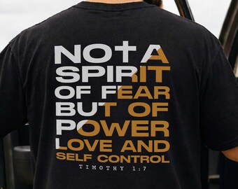 May include: Black t-shirt with white and gold text on the back. The text reads "NOT A SPIRIT OF FEAR BUT OF POWER LOVE AND SELF CONTROL TIMOTHY 1:7". The shirt is worn by a person standing next to a vehicle.