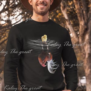 May include: A black sweatshirt with a graphic of a chicken and a chick. The graphic shows the chicken upside down in water with the chick standing on the water's surface. The text "Kaley The Great" is printed on the sweatshirt.