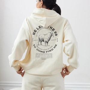 Pode incluir: Conjunto de moletom com capuz e calças de moletom creme. A parte de trás do moletom com capuz apresenta um gráfico circular com uma ilustração de ovelha e o texto "He left the 99 to come find me". O texto também inclui um versículo bíblico.