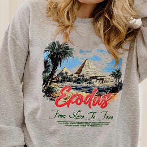 May include: A light gray sweatshirt featuring a colorful graphic of pyramids, palm trees, and the word "Exodus" in red script. Below the graphic are the words "From Slave To Free" in green script, with additional text below.