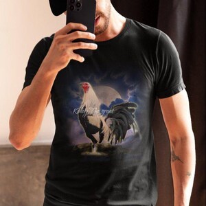 Puede incluir: Una camiseta negra con un estampado gr&aacute;fico de un gallo blanco y negro de pie sobre una roca con una luna llena en el fondo. El texto "Kiley the great" est&aacute; impreso en la camiseta.