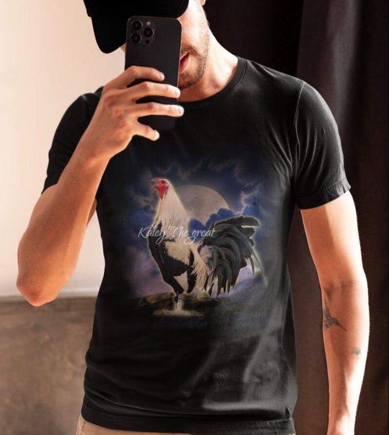 Puede incluir: Una camiseta negra con un estampado gr&aacute;fico de un gallo blanco y negro de pie sobre una roca con una luna llena en el fondo. El texto "Kiley the great" est&aacute; impreso en la camiseta.