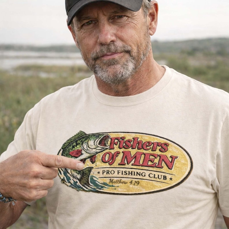 K&ouml;nnte beinhalten: Cremefarbenes T-Shirt mit einer Fischgrafik und dem Schriftzug "Fishers of Men Pro Fishing Club Matthew 4:19" in Rot und Gr&uuml;n. Eine Person zeigt auf das T-Shirt.