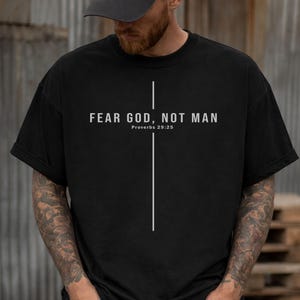 May include: Black t-shirt with the text "FEAR GOD, NOT MAN" and "Proverbs 29:25" in white. A vertical white line intersects a horizontal white line, forming a cross. The shirt has short sleeves and a crew neck.