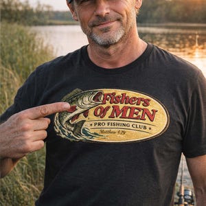 K&ouml;nnte beinhalten: Dunkelgraues T-Shirt mit einer Grafik, die einen Fisch zeigt, der aus dem Wasser springt, und dem Text "Fishers Of MEN PRO FISHING CLUB Matthew 4:19" in einem ovalen Design. Das T-Shirt wird von einer Person getragen, die auf die Grafik zeigt.