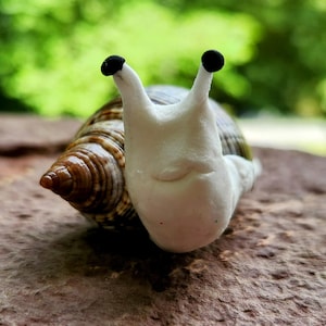 Può includere: Immagine dettagliata di una statuina a forma di lumaca. La lumaca ha un corpo bianco con due antenne nere. Il suo guscio è marrone con un motivo a strisce. La lumaca è appoggiata su una superficie marrone strutturata con uno sfondo verde sfocato.