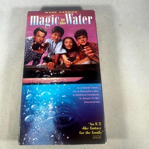 Puede incluir: Caja de VHS de la película "Magic in the Water" con Mark Harmon. La portada muestra un grupo de personas y el título "magic in the Water". La parte inferior presenta una escena submarina azul.