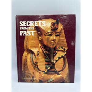 以下が含まれることがあります： 「Secrets From The Past」というタイトルのハードカバー本。表紙はバーガンディ色で、エジプトのファラオの金色の胸像が描かれています。National Geographic Societyによって出版されました。