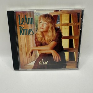 Puede incluir: Una funda de CD de LeAnn Rimes con una foto de la artista. La portada del álbum tiene el texto "LeAnn Rimes" en verde azulado y la palabra "blue" en una escritura azul oscuro. La funda del CD está en una funda de plástico transparente con lomo negro.