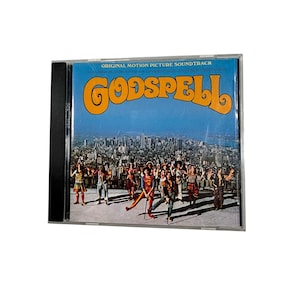 Puede incluir: Una caja de CD con la banda sonora original de "Godspell". La portada tiene un fondo de cielo azul con la palabra "Godspell" en naranja y un grupo de personas con trajes coloridos. La caja del CD está en una funda de plástico transparente.