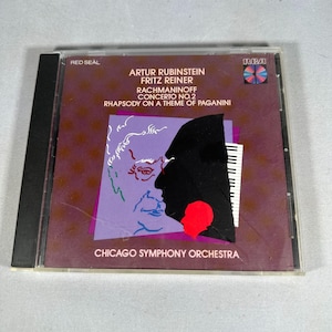 Puede incluir: Una caja de CD con una cubierta granate, que muestra texto y gráficos. El texto incluye "ARTUR RUBINSTEIN" y "RACHMANINOFF CONCERTO NO.2". La portada presenta una silueta de perfil y un diseño de teclado de piano. El CD está en una funda de plástico transparente.