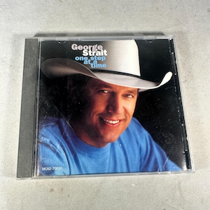Op de afbeelding: Een cd-hoesje met het album "One Step at a Time" van George Strait. De cover toont een lachende man met een witte cowboyhoed tegen een donkere achtergrond. De cd-hoes is van transparant plastic.