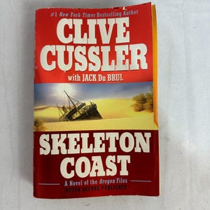 Może przedstawiać: Książka w miękkiej okładce zatytułowana "Skeleton Coast" autorstwa Clive'a Cusslera i Jacka Du Brula. Okładka przedstawia wrak statku w pustynnym krajobrazie. Tytuł i nazwisko autora są dużymi białymi literami na czerwonym tle.
