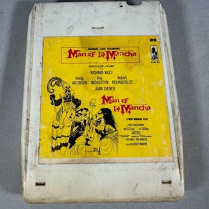 May include: A vintage 8-track tape cartridge for the musical "Man of La Mancha." The yellow label features the title in a stylized font, cast names, and a theatrical illustration. The cartridge is housed in a white plastic casing.