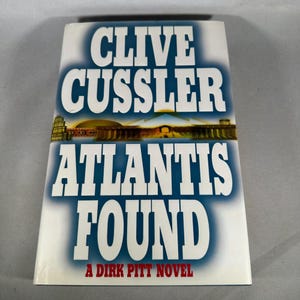 Könnte beinhalten: Ein Buch mit dem Titel "ATLANTIS FOUND" von Clive Cussler. Das Cover zeigt den Titel in großen weißen Buchstaben auf blauem Farbverlauf. Unter dem Titel befindet sich eine Grafik eines Gebäudes. Das Buch ist ein Dirk Pitt-Roman.