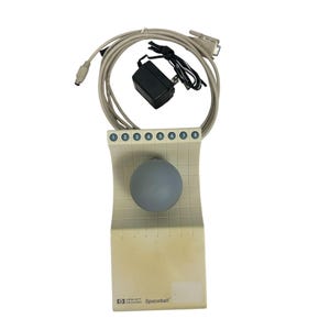Puede incluir: Un Spaceball Hewlett Packard beige con una bola gris, ocho botones numerados y un patrón de cuadrícula. Incluye un adaptador de corriente y un cable en espiral con conectores.