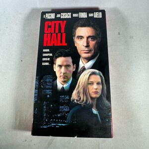 May include: A VHS tape for the movie "City Hall" featuring Al Pacino, John Cusack, Bridget Fonda, and Danny Aiello. The cover has a black background with the title in red and the actors' faces. The tape also lists "Murder, Corruption, Cover-up, Scandal."