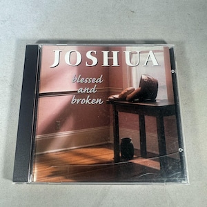 Pode incluir: Uma caixa de CD com o título "JOSHUA" em letras brancas. Abaixo, as palavras "blessed and broken" em itálico. A capa apresenta uma mesa com pão e uma cesta, e um vaso escuro.