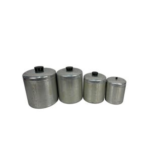 Puede incluir: Juego de cuatro botes de aluminio con tapas negras. Los botes son cilíndricos y varían en tamaño, con el más grande a la izquierda. Cada bote tiene texto impreso en el lateral.