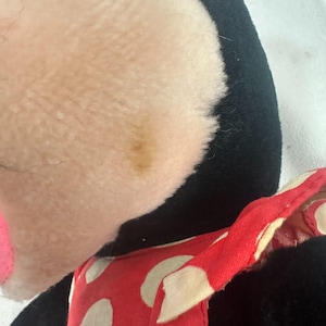 May include: Close-up of a plush toy with a black head and body and a light pink face. A red polka-dot bow is visible. The toy has a brown stain on its face.