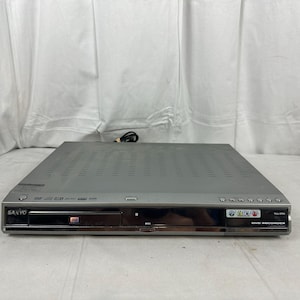 May include: A silver Sanyo DVD recorder featuring a front-loading disc tray with an RW logo, a central display panel, and a series of control buttons on the right side. The unit includes a label indicating AV inputs inside and a power button on the left.