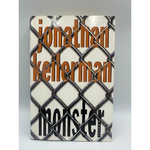 May include: A hardcover book titled "Monster" by Jonathan Kellerman. The book cover features the author's name in orange, the title in black, and a chain-link fence overlaying the text on a cream background.