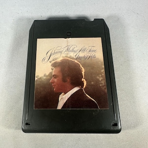 May include: Black 8-track tape featuring the album "Johnny Mathis All-Time Greatest Hits." The album cover shows a profile of Johnny Mathis in a suit. The text is in a cursive font.