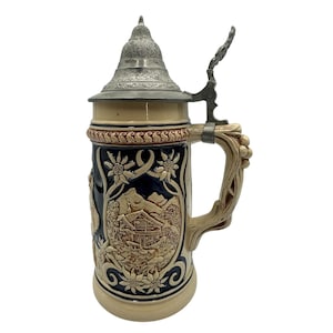 Puede incluir: Una jarra de cerveza de cerámica con un diseño en relieve detallado que presenta una escena de montaña, flores y una cabaña. La jarra tiene una tapa y un asa de metal, y es principalmente beige, azul y marrón.