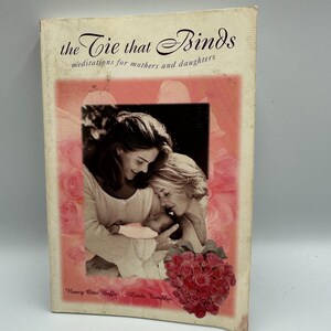 May include: A book titled "The Tie That Binds" with the subtitle "meditations for mothers and daughters." The cover features a photo of two women and a baby, with a bouquet of red roses at the bottom. The authors' names are at the bottom.