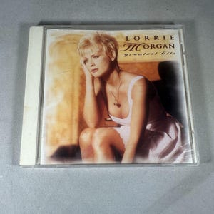 May include: A CD case featuring Lorrie Morgan's "greatest hits." The album cover shows a woman with short blonde hair, wearing a white dress and a necklace. The background has a warm, sepia tone.