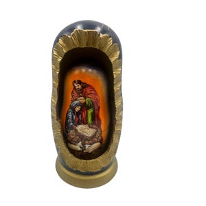 May include: A hand-painted wooden nesting doll depicting the Nativity scene. The exterior is dark blue with a gold-colored trim. Inside, a detailed painting shows Mary, Joseph, and baby Jesus in a manger, set against an orange background.