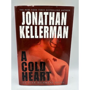 May include: A hardcover book titled "A Cold Heart" by Jonathan Kellerman. The cover is predominantly dark red with white text. The title and author's name are prominently displayed, with a blurred image of a person's back and arm.