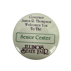 May include: A round, off-white button with text. The button reads "Governor James R. Thompson Welcomes You To The Senior Center" in purple text. Below, in a green rectangle, is "Senior Center". The bottom of the button reads "ILLINOIS STATE FAIR" in purple.