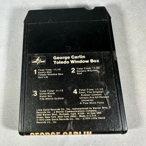Puede incluir: Una cinta de 8 pistas negra del álbum de comedia "Toledo Window Box" de George Carlin. La etiqueta incluye títulos de canciones como "Goofy Shit" y "The Metric System". Se ve el logotipo de Little David Records.