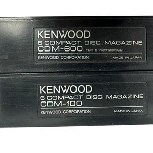 May include: Two black Kenwood 6 compact disc magazines, models CDM-600 and CDM-100. Each unit features white text labels reading Kenwood Corporation, Made in Japan, and specifies compatibility for 5-inch CDs. The rectangular plastic cases are stacked.