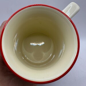 May include: A ceramic mug with a cream-colored interior and a red rim. The handle is white. The mug is viewed from above, showcasing the inside of the cup. The mug is likely used for drinking beverages.