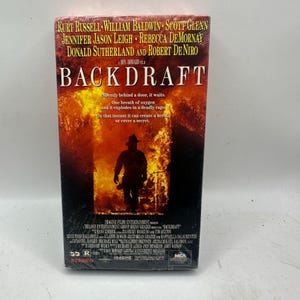 Peut inclure: Une cassette VHS du film "Backdraft" avec Kurt Russell, William Baldwin et Robert De Niro. La jaquette montre la silhouette d'un pompier devant un fond de flammes, avec le titre en lettres grasses.