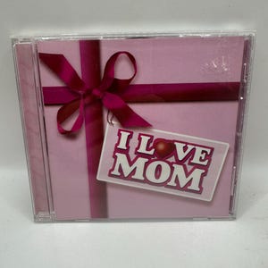 Puede incluir: Una caja de CD con un fondo rosa y un diseño de lazo rosa oscuro. Las palabras "I Love Mom" están impresas en una etiqueta blanca con un corazón rojo.