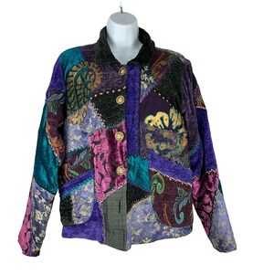 May include: A long-sleeve jacket featuring a patchwork design with various textures and patterns in shades of purple, teal, black, and gold. The jacket has a dark collar, a front button closure with four gold-toned buttons, and two front pockets.
