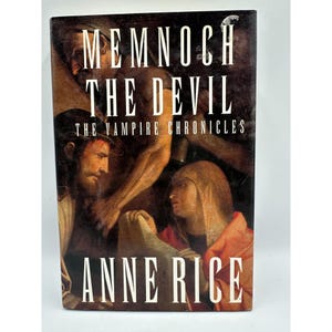 May include: A hardcover book titled "Memnoch the Devil" by Anne Rice. The cover features a dark, artistic illustration with figures in shades of brown, red, and white. The book is part of "The Vampire Chronicles" series.