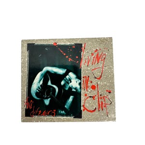 May include: Album cover for Ani DiFranco's "Living in Clip." The cover features a black and white image of a person playing an acoustic guitar. The title is in red, with red splatter accents.
