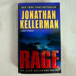 May include: A paperback book titled "Rage" by Jonathan Kellerman. The cover features the title in large red letters, the author's name in gold, and a dark blue background with a car. The book is labeled as a New York Times Bestseller.