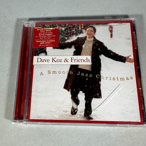 Könnte beinhalten: Eine CD-Hülle für "Dave Koz & Friends: A Smooth Jazz Christmas". Das Cover zeigt einen Mann in einem braunen Mantel in einer verschneiten Szene. Die CD-Hülle hat einen roten Rand und enthält Text über besondere Gäste und Weihnachtsgeschenke.