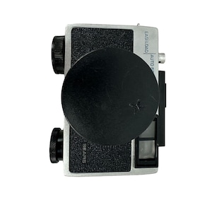 May include: A vintage Sears Easi-Load camera with a black lens cap. The camera body is silver and black, with the word "SEARS" printed on the front. The lens cap has a star emblem.