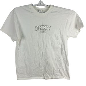 May include: White DKNY USA t-shirt. The short-sleeved shirt has a crew neck and the DKNY logo in a brown font. The shirt is hanging on a white plastic hanger against a white background.