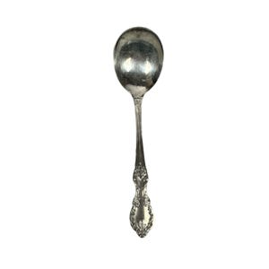 Puede incluir: Una cuchara de metal plateado con un cuenco redondeado y pulido junto a un mango decorativo. El mango presenta intrincados patrones florales y de volutas en relieve que se extienden desde el cuello hasta la base redondeada y ensanchada.