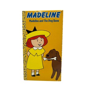 May include: A yellow book cover featuring the title "MADELINE" in blue letters. The cover shows a cartoon illustration of a girl in a yellow dress and hat, and a brown dog holding a bone. The text "Madeline and The Dog Show" is also visible.