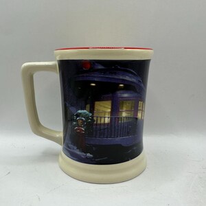 May include: A ceramic mug with a cream-colored base and handle, a red rim, and a dark blue exterior. The mug features a winter scene with a train car and falling snow. The handle is rectangular.