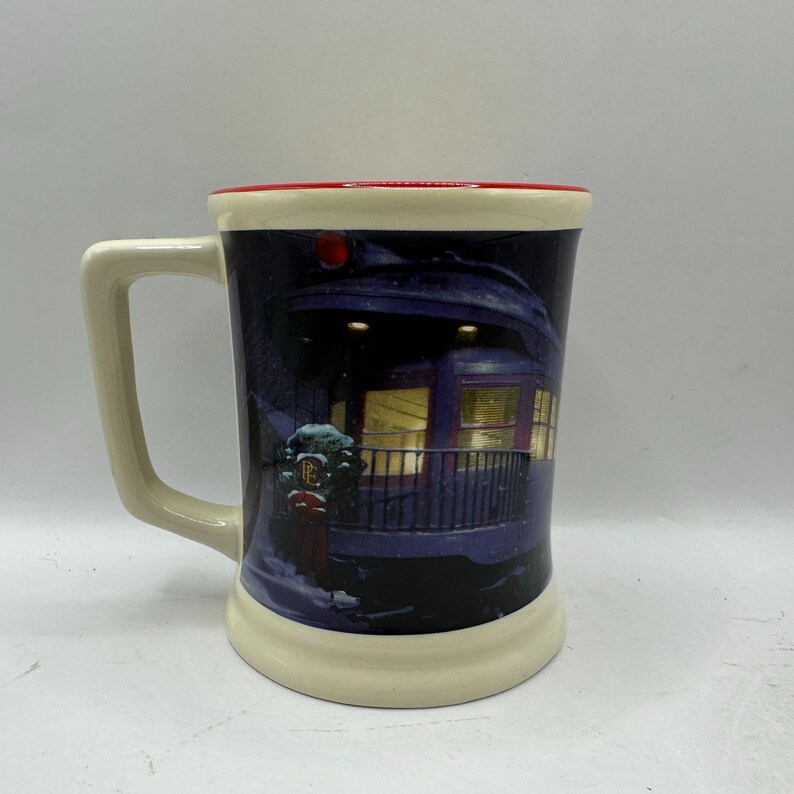 May include: A ceramic mug with a cream-colored base and handle, a red rim, and a dark blue exterior. The mug features a winter scene with a train car and falling snow. The handle is rectangular.