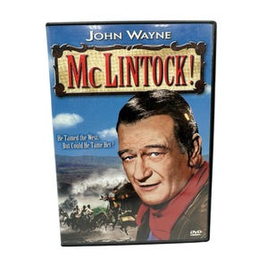 Puede incluir: DVD de la película "McLintock!" protagonizada por John Wayne. La portada presenta un retrato de Wayne, con el título en letras grandes y ornamentadas. El fondo muestra un paisaje del Oeste y un grupo de personas a caballo.